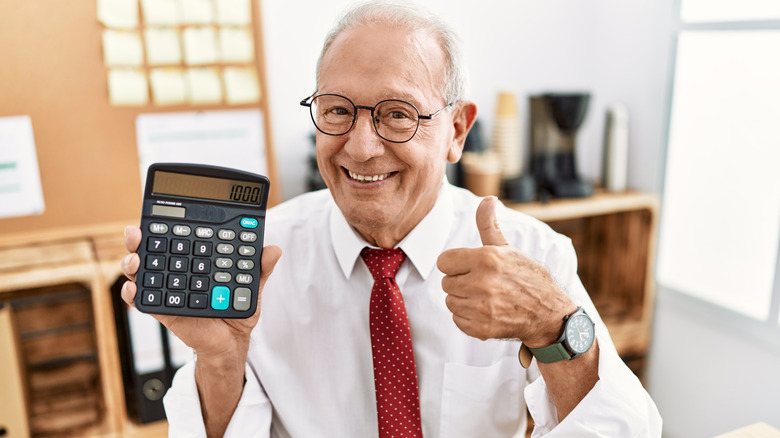 A smiling senior man sitting at a desk giving a thumbs up sign of approval while holding up a calculator.