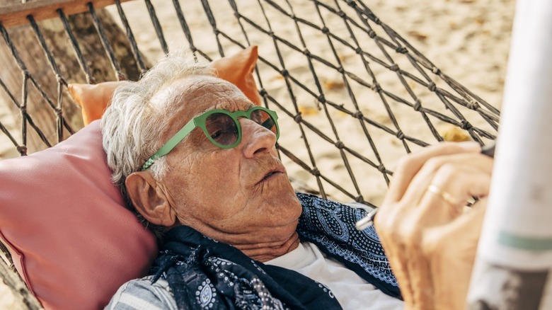 elderly caucasion man wearing green glasses laying on a hammock and a pink and orange pillow looking down at a booklet
