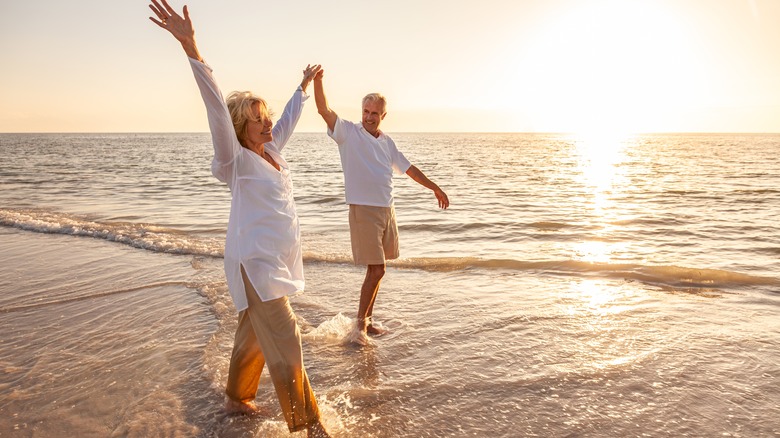 Happy senior man and woman holding hands with arms raised on a beach at sunset