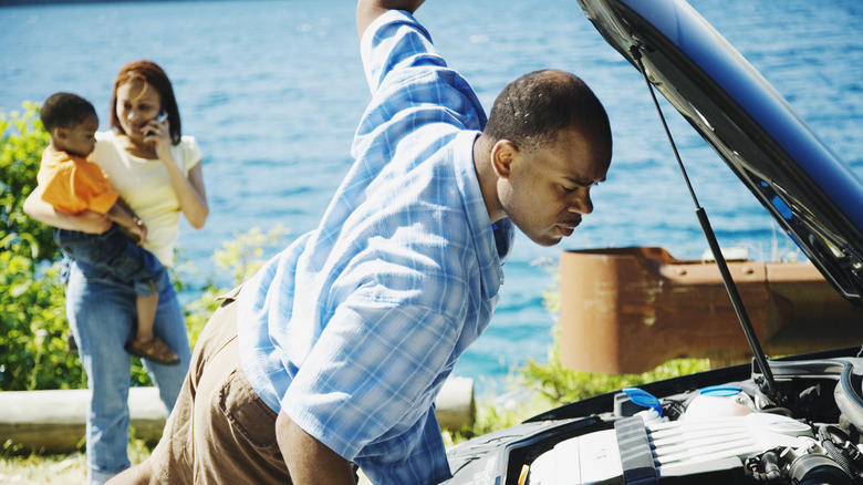 A man peers under the hood of a car while a woman holding a child is making a phone call in the background.