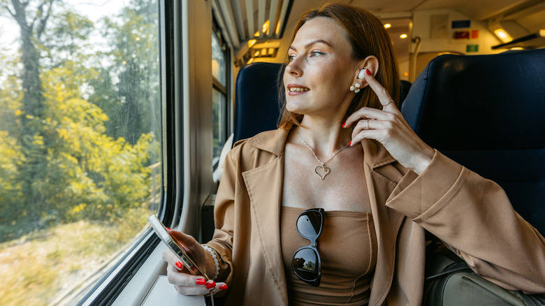 A Gen X woman with earbuds holding her phone while gazing out the window of a moving train.