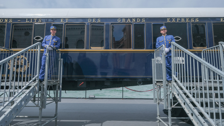 Two train porters at the top of stairs in front of a luxury train in Europe.