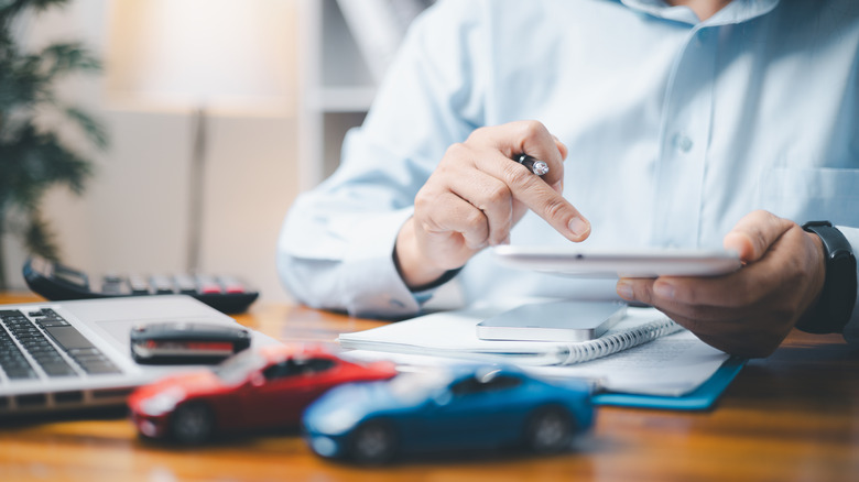 Faceless man with tablet calculator sits at desk with two model cars on it as he figures out expenses.