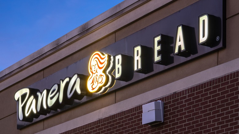 A lightly burnt out Panera Bread sign over a brick storefront, against a twilit sky