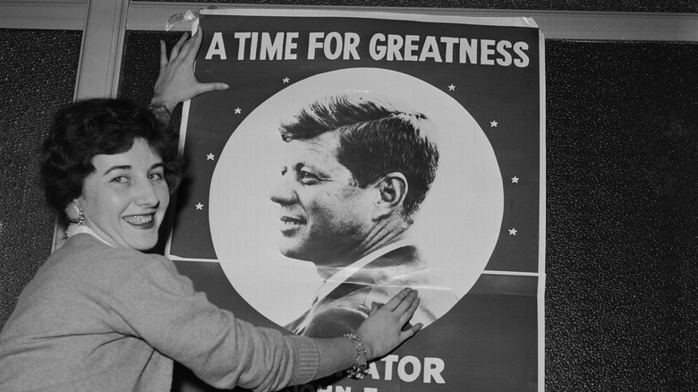 Black and white image of woman posing in front of a John F. Kennedy campaign poster