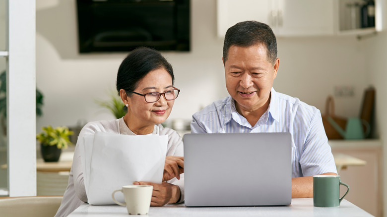 An elderly couple sit at a desk with an open laptop computer, paperwork, and calculator as they balance their bills in a sunny room