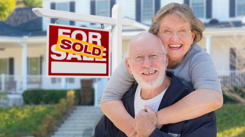 A smiling senior couple standing in front of a home with a sold sign in front.
