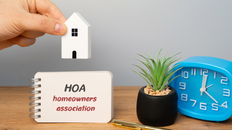 Hand holds small toy house over notebook with "HOA homeowners association" next to plant and clock