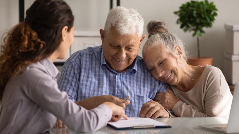A happy senior couple embracing while signing the paperwork to buy a home with a female realtor in front of them.