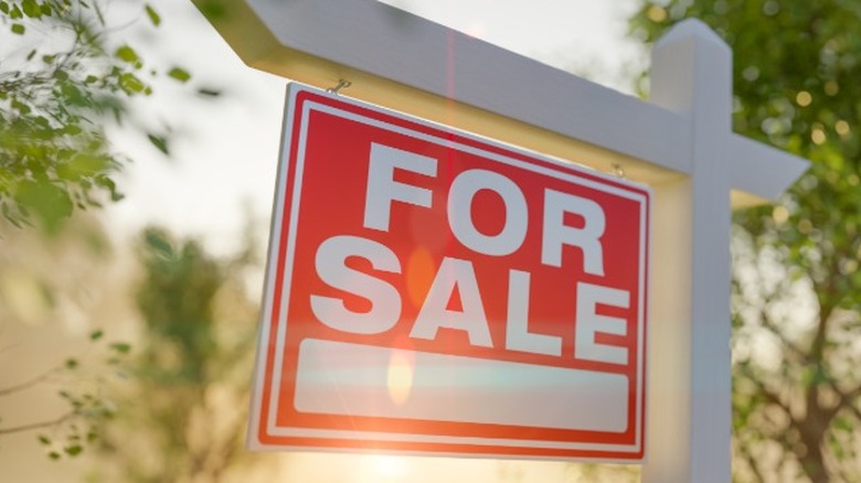 A red For Sale sign hanging from a white wood post with sunlight shining on it.