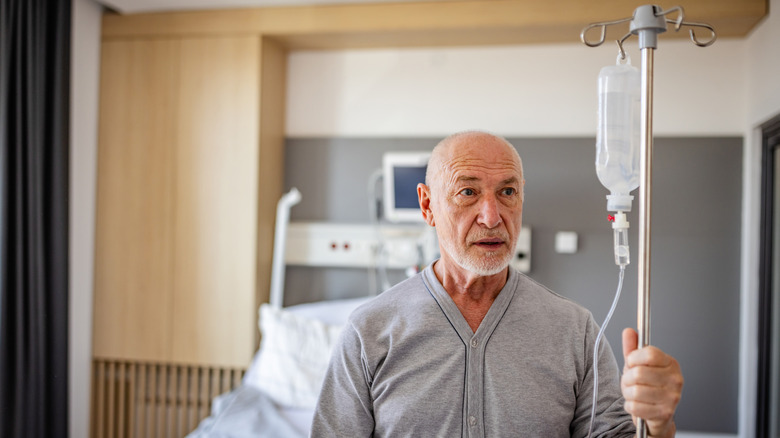 A retired man with a worried expression in a hospital room with his hand on an IV pole.