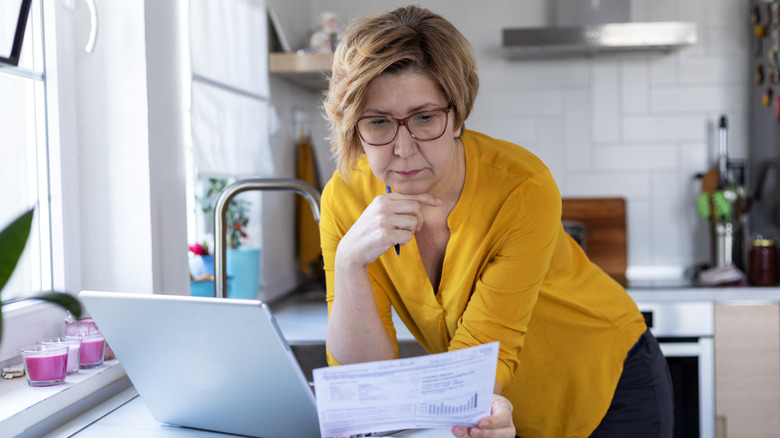 A female retiree standing in the kitchen looking at paperwork with an open laptop on the counter.