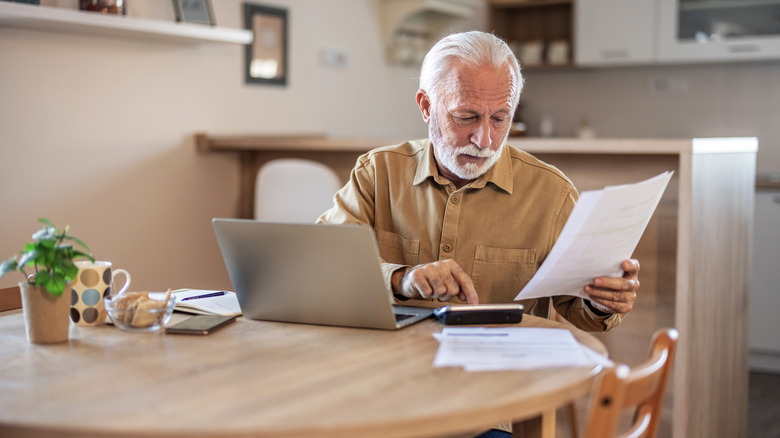 Retired man calculating something with a document in his hand and a laptop open