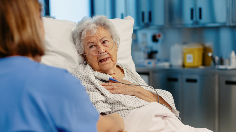 Old woman with nose tube sits on reclining bed in hospital while talking to healthcare professional
