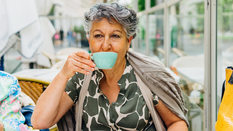 A retired woman drinking coffee in an outdoor cafeteria.