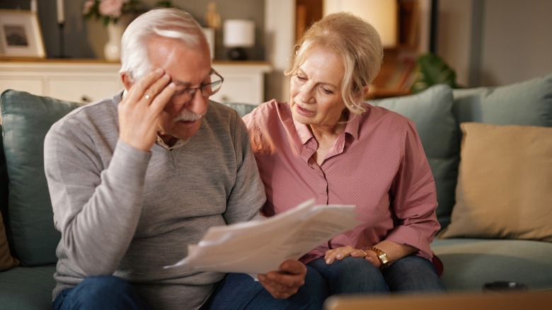 Older couple reviewing paperwork on a sofa, with the man resting a hand against his forehead