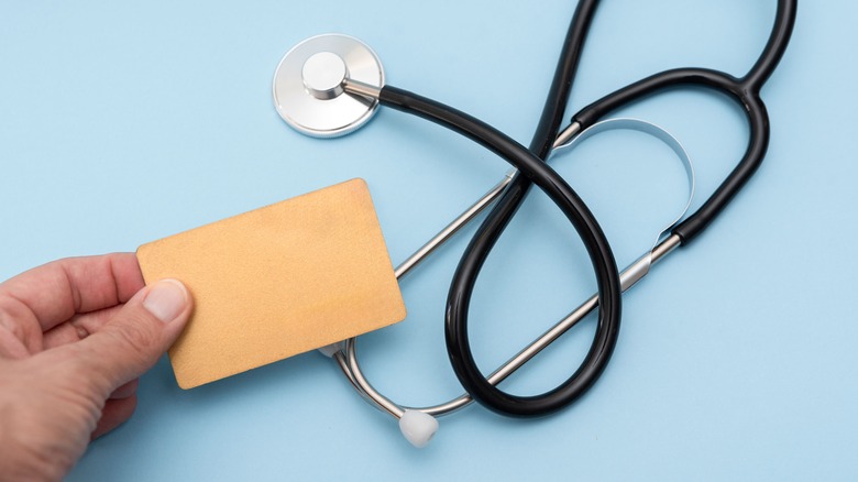 A close up of hand holding blank insurance card next to stethoscope on blue background