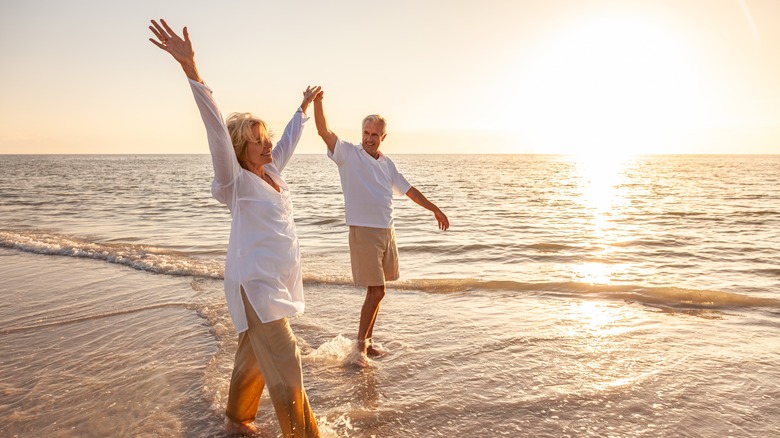 Happy senior man and woman old retired couple walking and holding hands arms raised on a beach at sunset.
