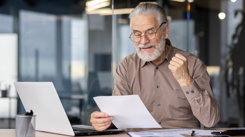 Senior businessman excitedly celebrating success while reviewing documents on laptop in modern office.