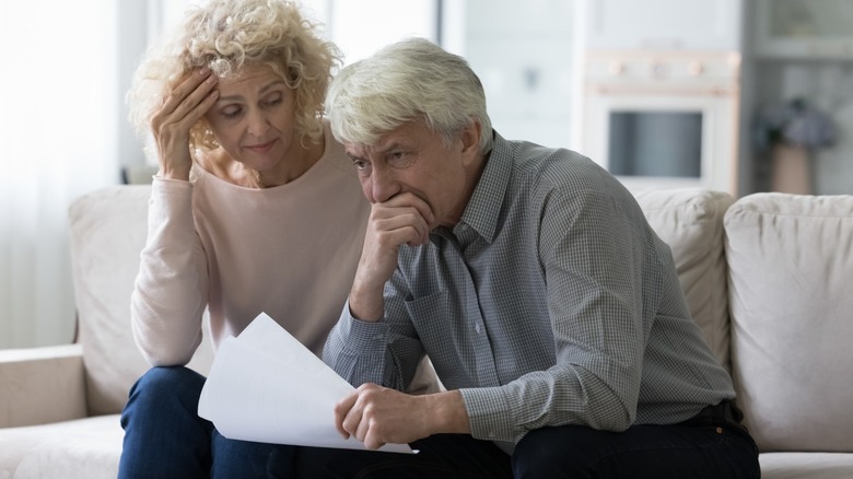 An elderly couple looking concerned reading paperwork in their living room.