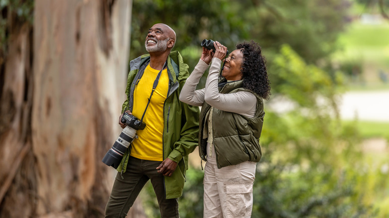 An older couple smiling, hiking, and taking in the sights with binoculars and a camera