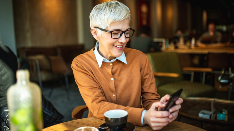 A smartly dressed senior woman smiling while perusing her phone in a cafe.