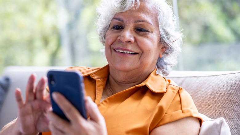 Smiling older woman seated on her couch as she looks down at her smartphone