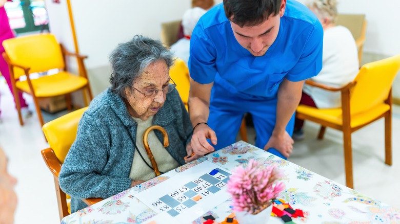 Nurse helping an elderly woman playing bingo, providing care and support in a nursing home environment.