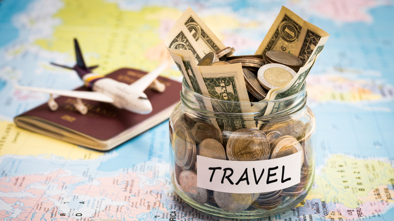 A jar of money labeled Travel sitting on a map with an airplane on a passport in the background.