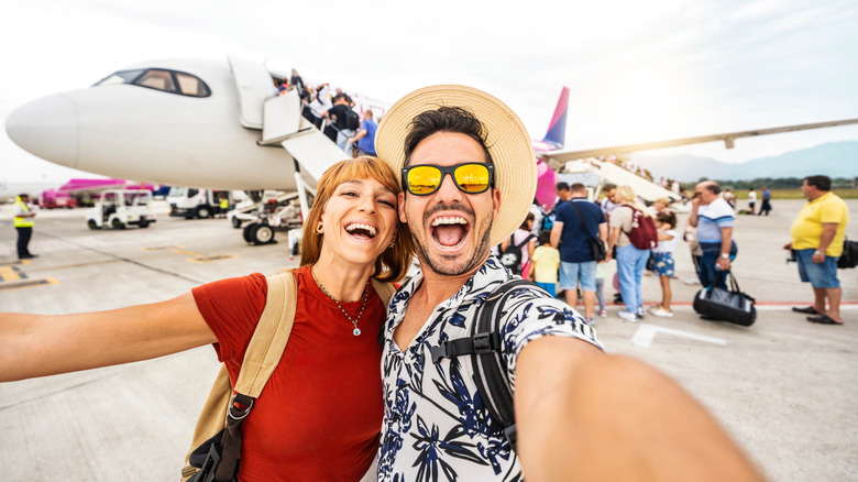 A happy couple taking a selfie in front of an airplane with people in line to board in the background.