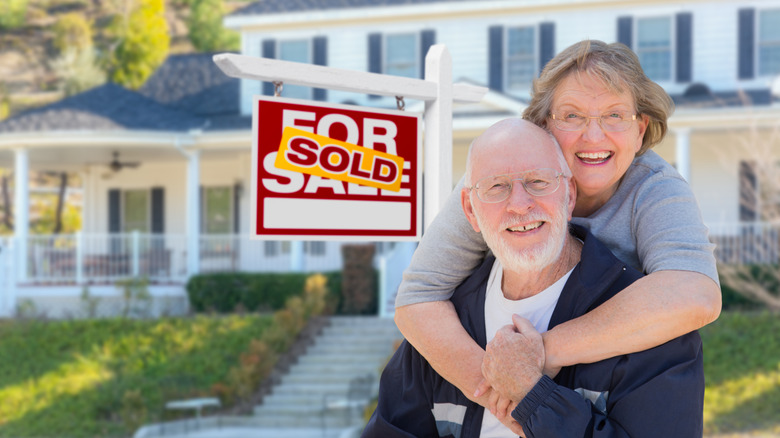 Older couple in front of house for sale sign
