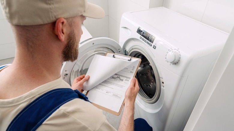 A technician installing a new washing machine and checking paperwork for it on a clipboard.