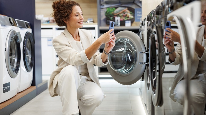 A woman in an appliance store shopping for washers and dryers kneeling in front of a washer with the door open with her phone out.