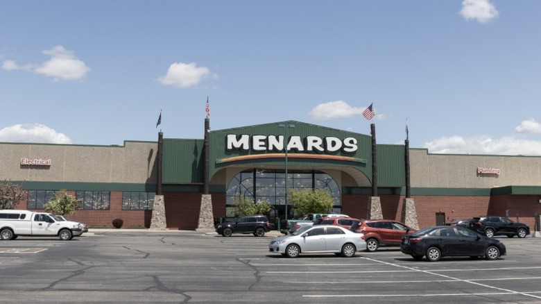 The exterior of a Menards store in the United States with a large parking lot outside of it dotted with vehicles.