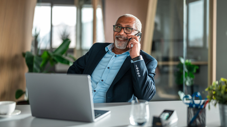 older man sitting in an office on the phone with a laptop open in front of him
