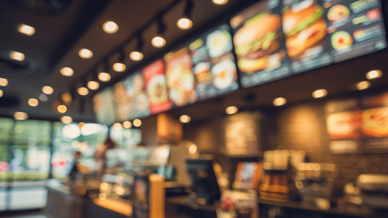 A blurred image of a fast-food restaurant's interior, showcasing the counter, menu boards displaying various food items, and a few indistinct customers.