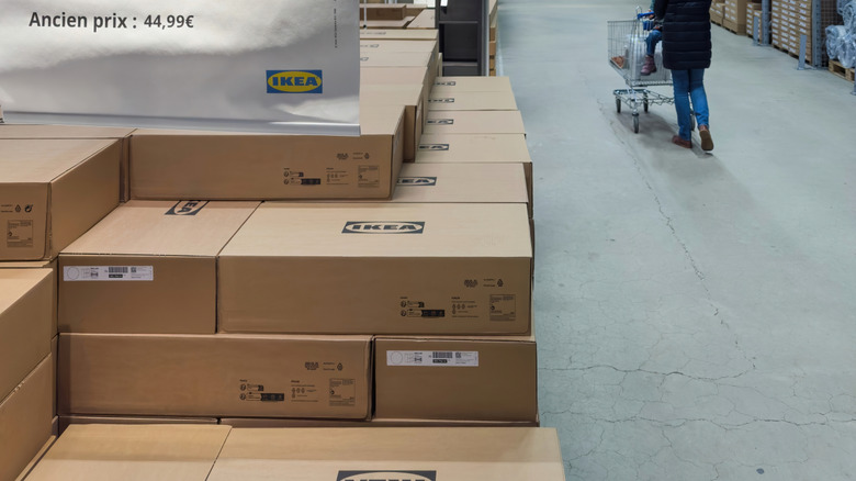 Interior of IKEA warehouse section showing stacked boxes of KALLAX shelving units with a large price sign and a customer pushing a shopping cart along the aisle.