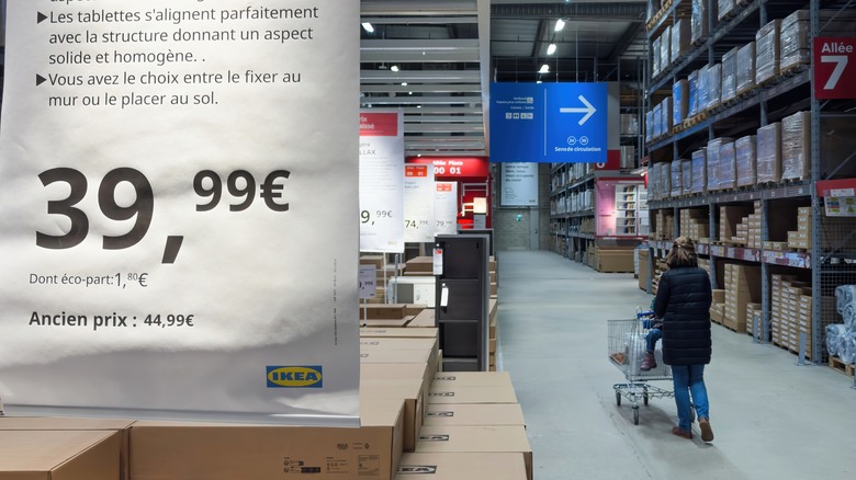 Interior of IKEA warehouse section showing stacked boxes of KALLAX shelving units with a large price sign and a customer pushing a shopping cart along the aisle.