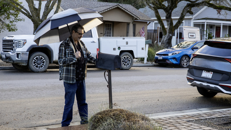 A smiling older man holding an umbrella checks his mailbox on a suburban street