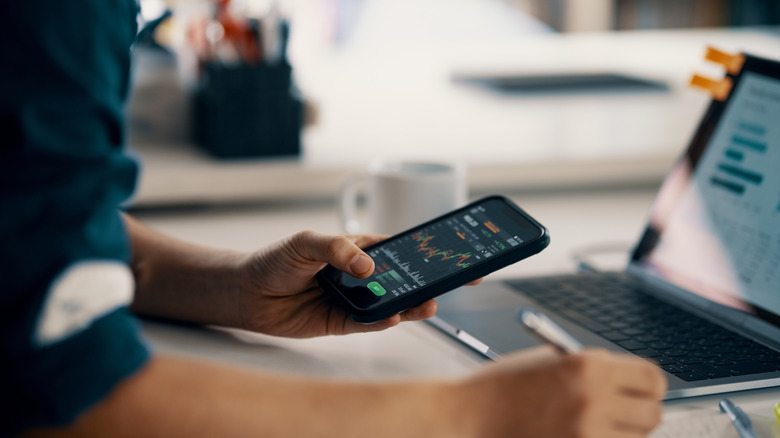 An investor doing calculations with a pen with stock information displayed on a phone and computer