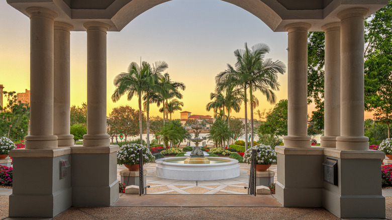 A garden with a fountain and palm trees through the view of a stone archway