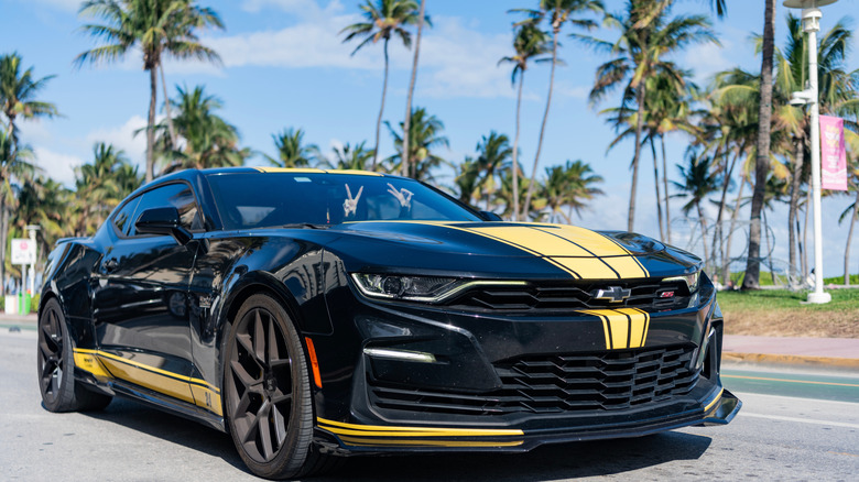 A Chevrolet Camaro parked on a palm tree lined street with an anonymous driver flashing a peace sign behind the wheel.