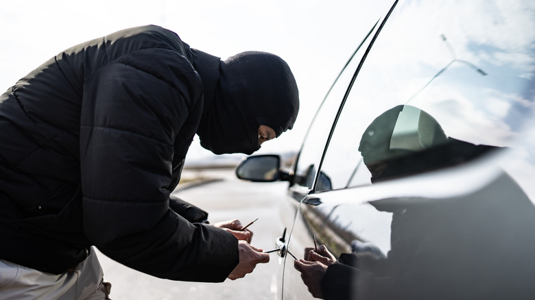 Masked individual in dark clothing trying to pick the lock of a parked car.