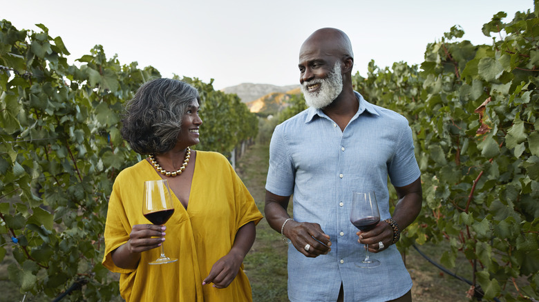 An elegant mature couple walking through a vineyard with glasses of wine.