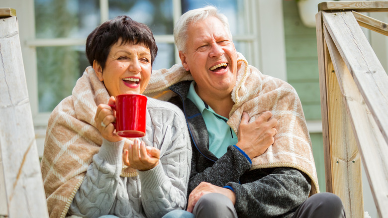 A laughing mature couple huddled together under a blanket on their front porch
