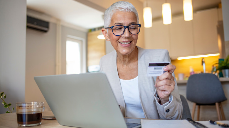 A smiling mature woman holding her credit card in front of a laptop while paying bills.