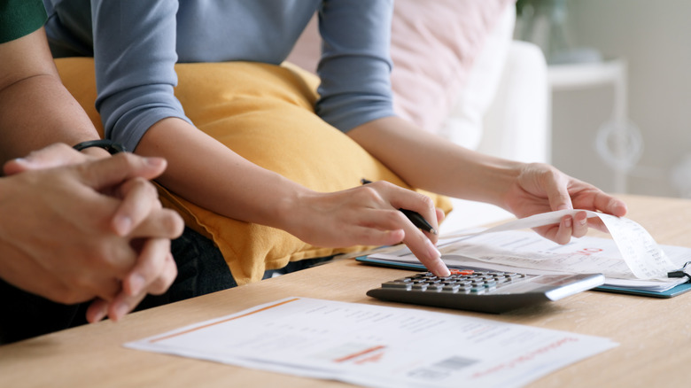 Woman with pillow in lap uses calculator while man clasping hands sits beside her