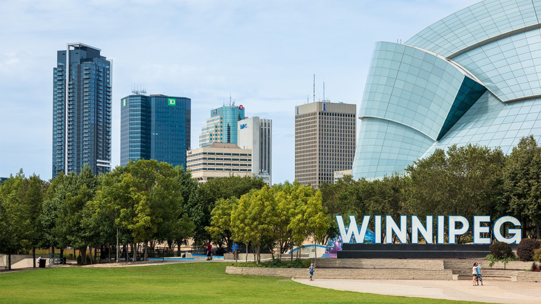 Winnipeg sign in front of trees with skyscrapers in the distance