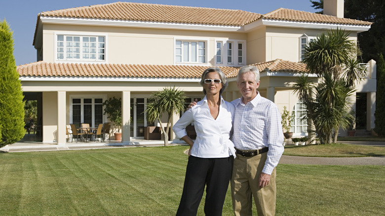 A mature couple standing on a large manicured lawn and a big house.