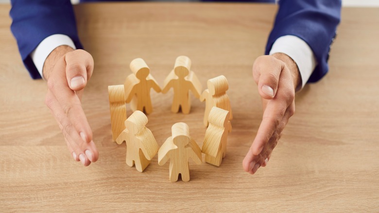 A circle of wooden figures flanked by a man's hands to symbolize employer-based health insurance.